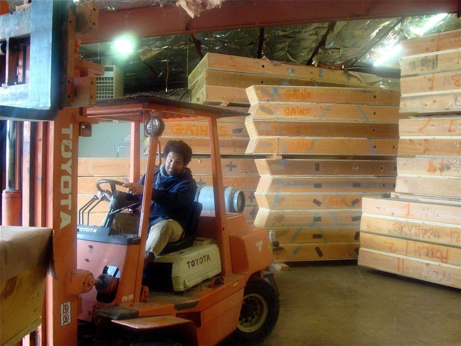 Orange forklift carrying stacked wooden panels in a warehouse, with a person driving inside the cab