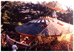 Round wooden house with a conical roof in a wooded area, viewed from above at sunset