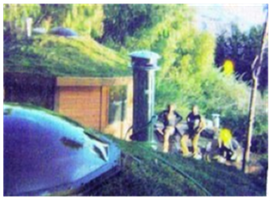 People standing by a tall column near a grassy hillside and a house, with a car in the foreground