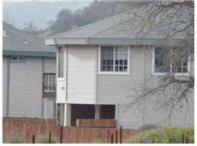 Two-story beige house with raised lower level, gray roof, and a wooden fence in front