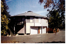 Round beige house with a dark roof, exterior stairs, and garage doors on a sunny hillside.