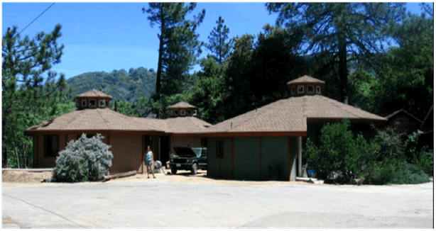 Single-story brown building with a wide roof, trees, and mountains in the background on a sunny day