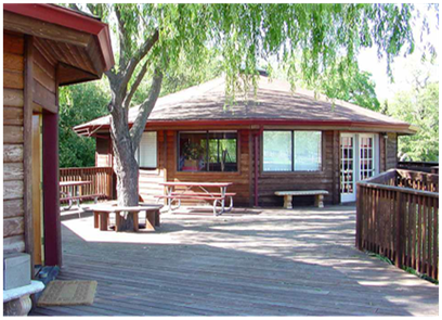 Wooden cabin deck with picnic tables and benches under leafy trees, viewed on a sunny day