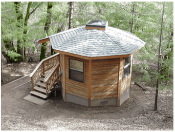 Small octagonal wood cabin with a shingled roof and stairs, set among trees in a forest.