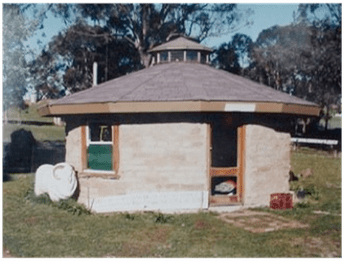 Round brick house with a low, sloped roof, green window, and a small porch in a grassy yard