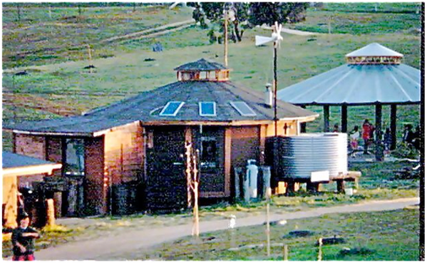 Rural homestead with round metal water tank, small house, and a thatched gazebo on a grassy hillside