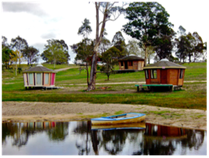 Small colorful huts by a pond, with trees and reflections in a grassy park.