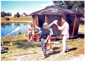 People feeding a white bird beside a small wooden cabin by a lake