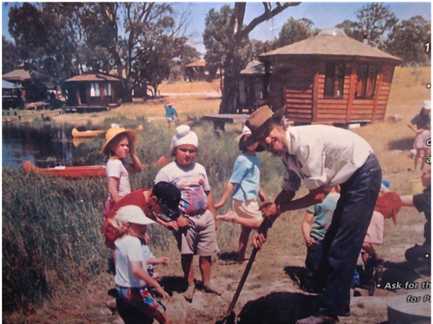 Children gardening with an adult beside a wooden house in a sunny yard