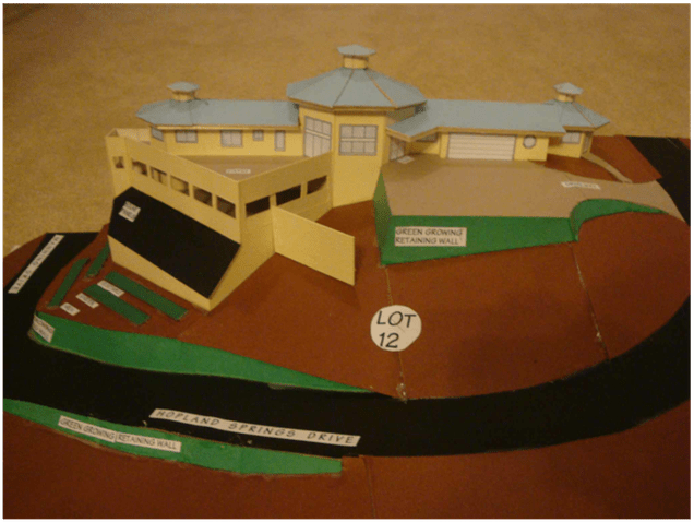Cardboard model of a yellow house on a landscaped lot with a black road and green lawn, marked “Lot 12”