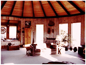 Rustic living room with a round wood-beamed ceiling, red walls, large windows, and wicker chairs