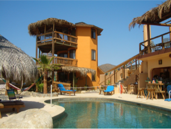Tropical resort pool with thatched-roof buildings, lounge chairs, and a bright blue sky