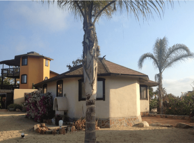 Single-story house with tan walls and a palm tree in a dry yard under a blue sky.