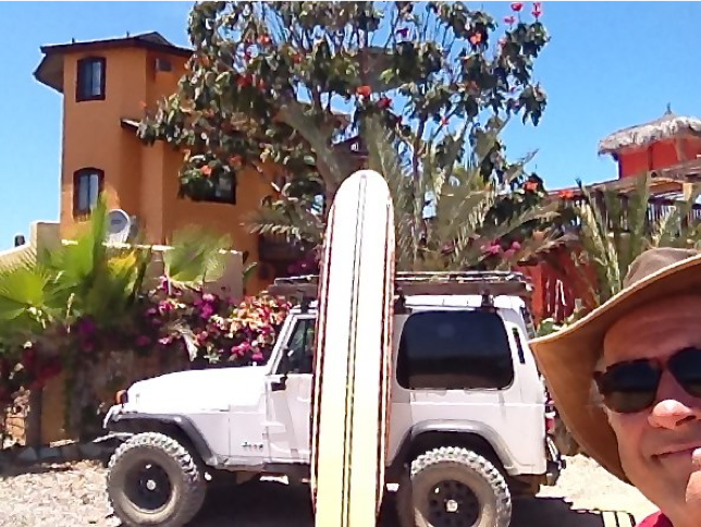 Man in a sunhat beside a white SUV with surfboards on the roof, tropical buildings and palm trees behind.