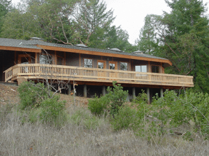 Wooden house with a long deck on a hillside, surrounded by trees and brush