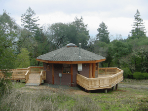 Small round wooden cabin with a wraparound deck in a wooded clearing