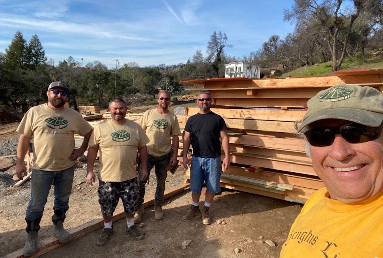 Five people posing beside stacked lumber outdoors on a sunny day.