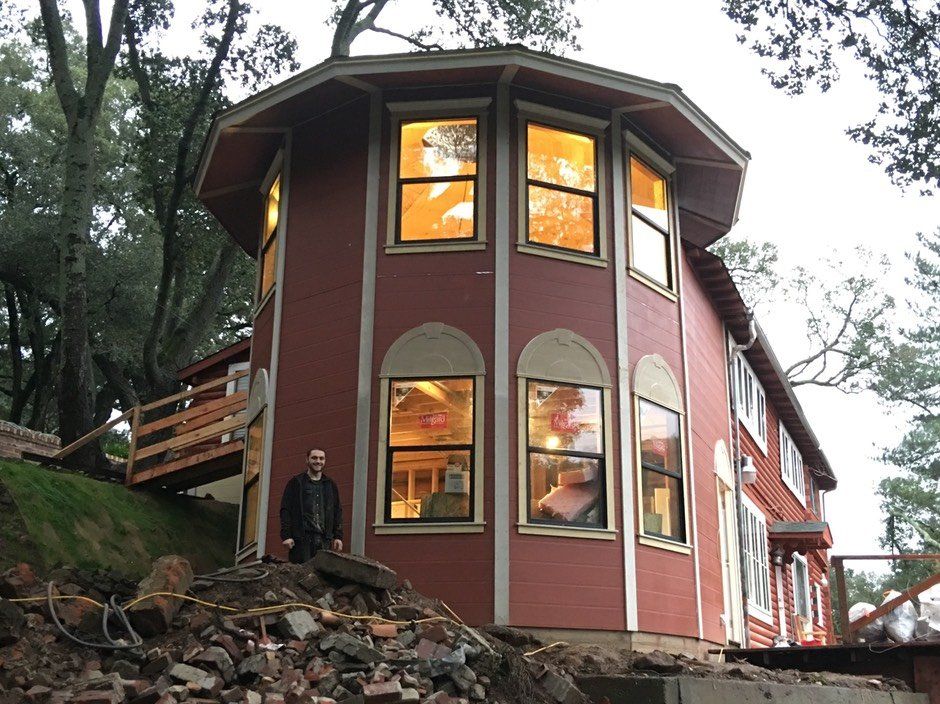Two-story red house with large windows on a wooded hillside, viewed from below