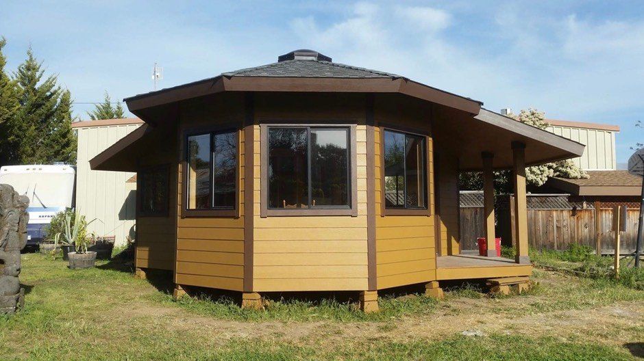 Small brown octagonal house with large windows and a covered porch in a grassy yard