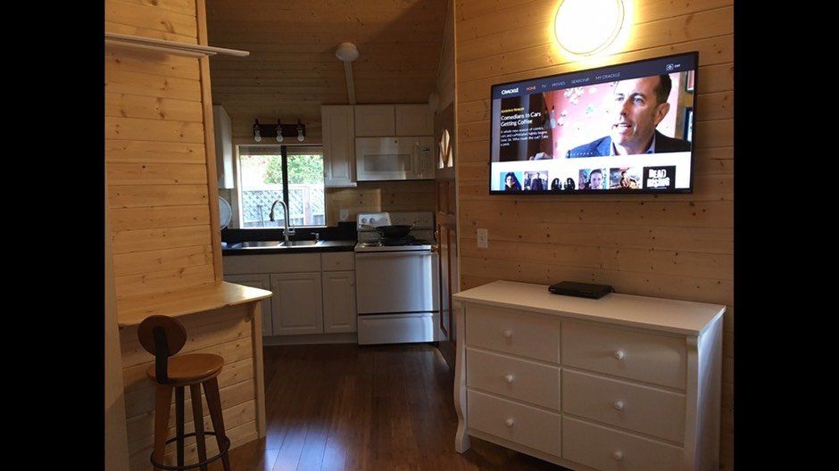 Cozy wood-paneled room with wall-mounted TV, white dresser, small kitchen, and a stool by the window.