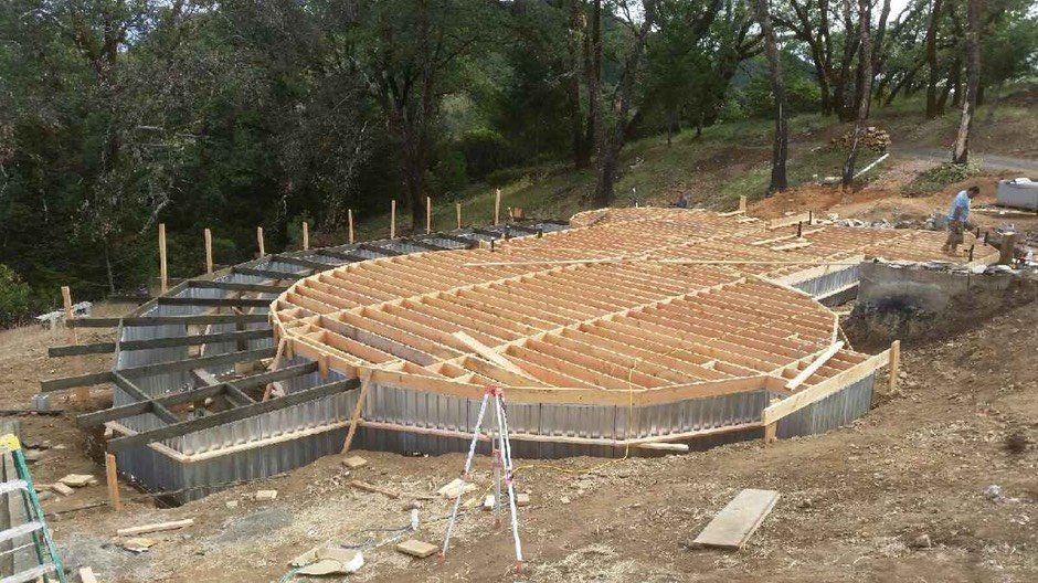 Circular wooden foundation under construction on a wooded hillside, with a ladder and formwork.