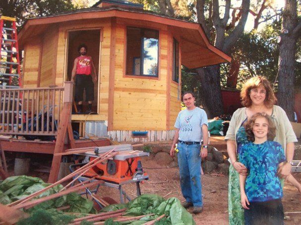 Three people stand by a small wooden cabin under construction, with a person on the porch and lumber in front.
