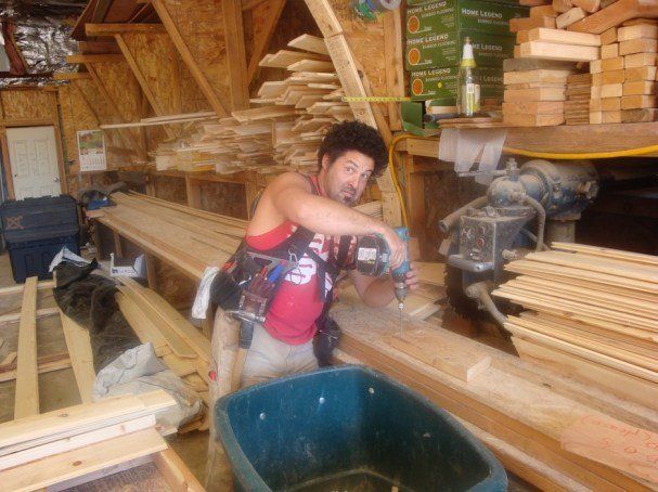 Person using a saw in a wood shop, with stacked lumber and tools around them.