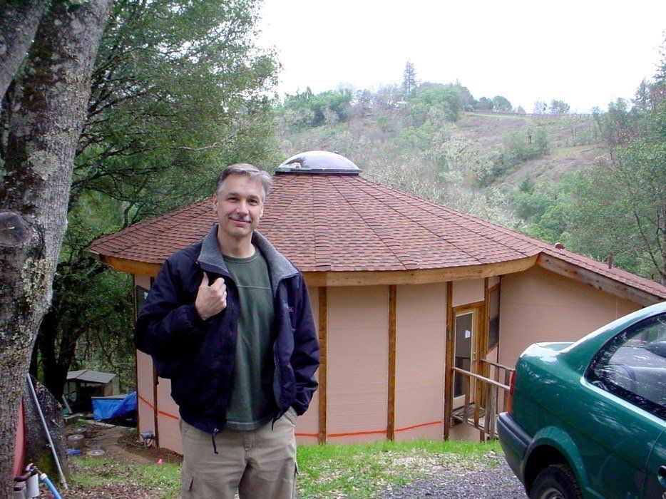 Man standing by a small round cabin in a wooded hillside, with a green car in the foreground.