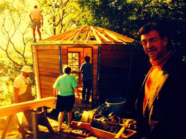 People building a small wooden cabin outdoors, with a man in the foreground and tools nearby.