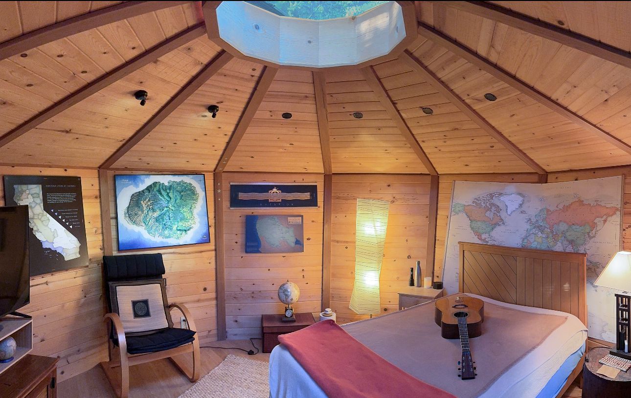 Cozy wooden yurt interior with a bed, chair, window map, and skylight above.