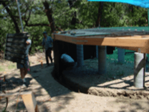 Outdoor shelter with a green roof and stone pillars in a wooded area, with two people nearby