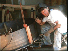 Person in a workshop leaning beside a wooden boat under construction, waving at the camera.