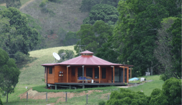Round wooden cabin with a red roof on a green hillside, surrounded by trees and misty hills.