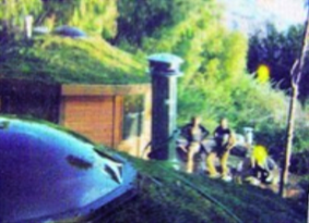 People near a house with a green roof; a blue car is in the foreground.