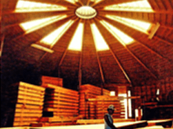 Stacked lumber inside a circular wooden warehouse under a bright skylight; a person stands below.