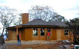 People standing outside a round wooden building under construction with windows and a brick chimney