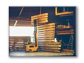 Forklift lifting stacked wooden pallets in a warehouse, with more pallets and a ladder nearby.