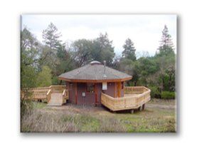 Small octagonal wooden cabin with wraparound deck in a wooded clearing under a cloudy sky