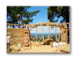 Outdoor hay-bale structure under construction with ladder, wooden frame, and ocean view in the background