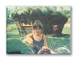 Woman in a tank top standing outdoors near a large planter or vehicle, with greenery in the background