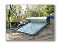 Roof-mounted solar water heater with blue panels on a shingled house in a wooded yard