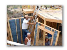 Workers building a wooden structure, with one man on a ladder inside a framed wall.