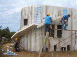 Workers plastering the exterior of a curved building under construction, with scaffolding and ladders.
