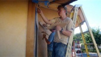 Person working on the side of a house, painting the eaves from a ladder.