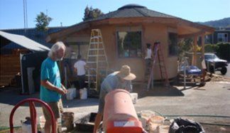 Two people working outside a peach-colored house, with ladders and a red cement mixer in the yard.