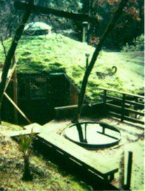 Rustic outdoor water wheel beside a small green pond and wooden platform under trees