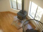 Cozy corner with a black wood stove, light rug, wooden floor, and chair by a sunlit window.