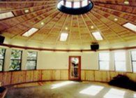 Round wooden room with windows, sunlit floor, and a central doorway under a domed ceiling