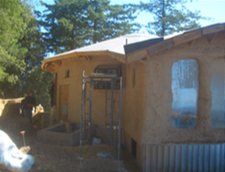 Small adobe-style house with a porch, trees behind it, and a corrugated metal fence in front.