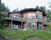 Stone house with wooden deck on a grassy hillside, surrounded by trees and large windows